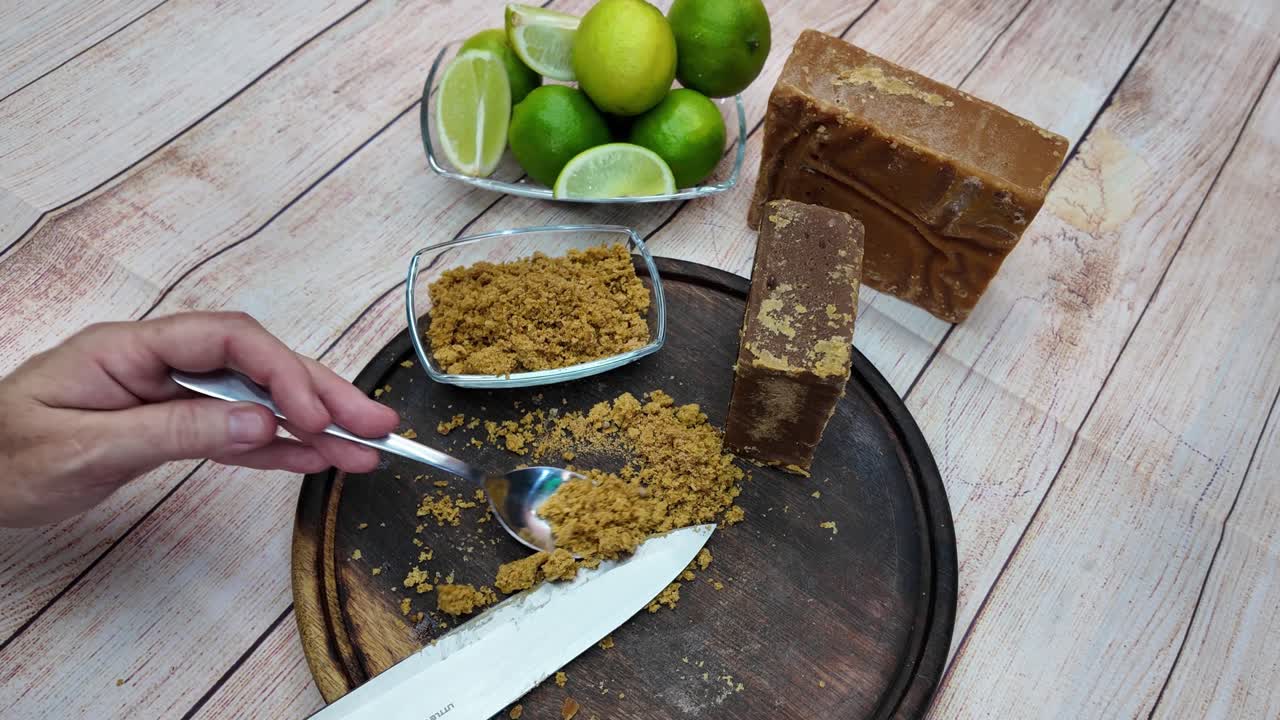 Close-up shot hand scooping and measuring grated panela (papelón) with a spoon, ingredients: raw sugar blocks and the fresh green limes, ready for traditional papelón con limón beverage