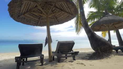 Vista-De-Una-Tumbona-Vacía-Cerca-De-Sombrillas-Nativas-Y-Palmeras-Contra-El-Agua-Azul-De-La-Isla-Mauricio