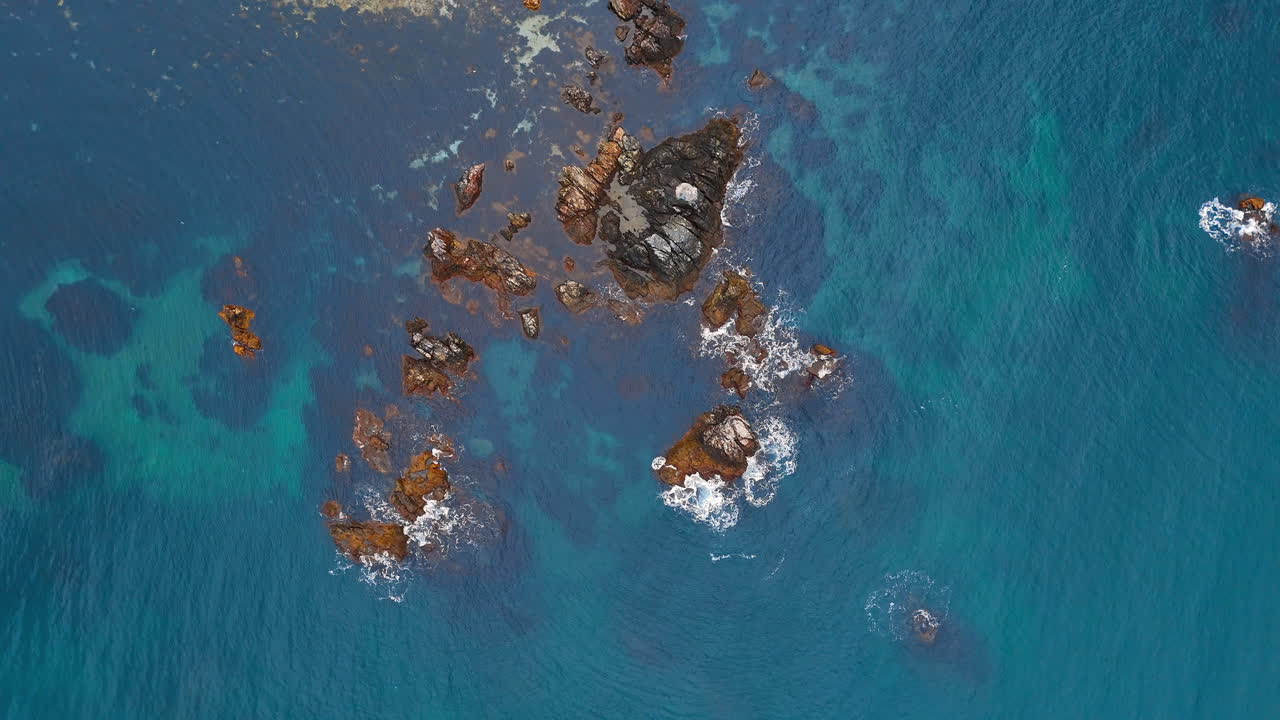 Aerial top down view of jagged sea rocks in blue water. Dark volcanic rocks surrounded by clear turquoise and deep blue ocean water
