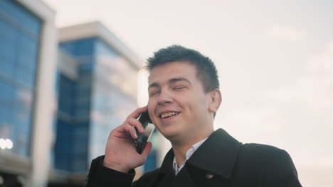 man in black coat happily talking on phone with excitement while making hand gesture in vibrant urban area with modern glass buildings, soft sunlight, and cheerful atmosphere