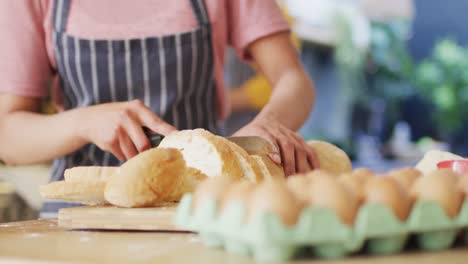 Video-of-midsection-of-biracial-woman-in-apron-slicing-bread-in-kitchen,-with-copy-space