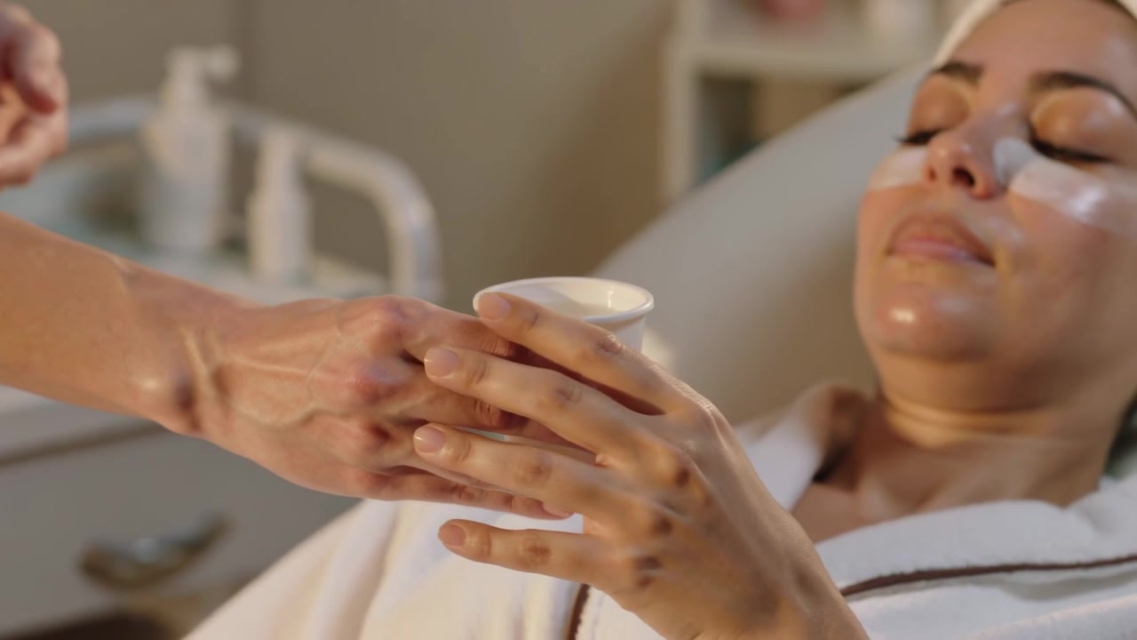 Soignant en tenue médicale tendant une tasse, un client allongé l'acceptant pendant un soin du visage avec des patchs pour les yeux