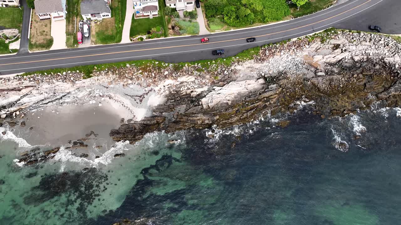 Drone view of cars driving along scenic road as waves crash on rocky coast