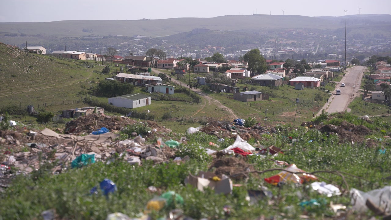 Household waste covers a grassy slope in the foreground while small homes and a paved road stretch across the valley beyond.