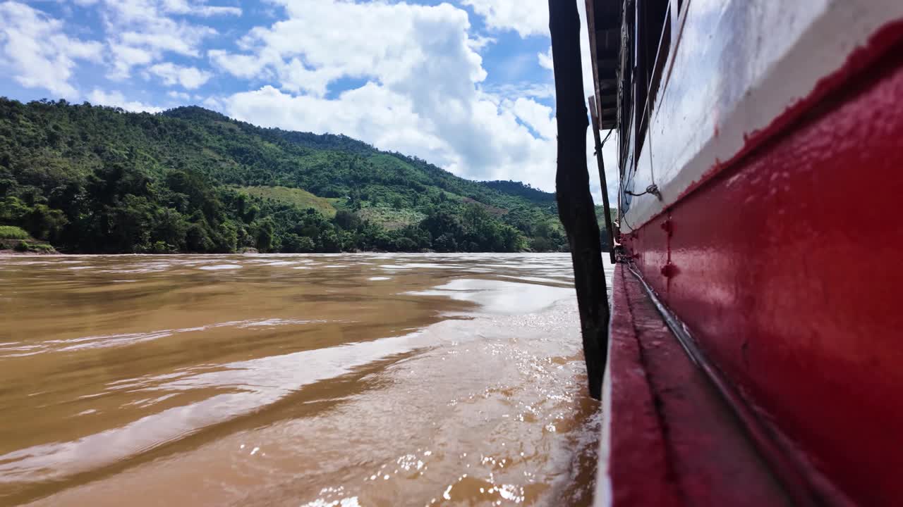 A slow boat glides along the Mekong in Laos, passing lush riverbank and tropical jungle. Low Angle Shot