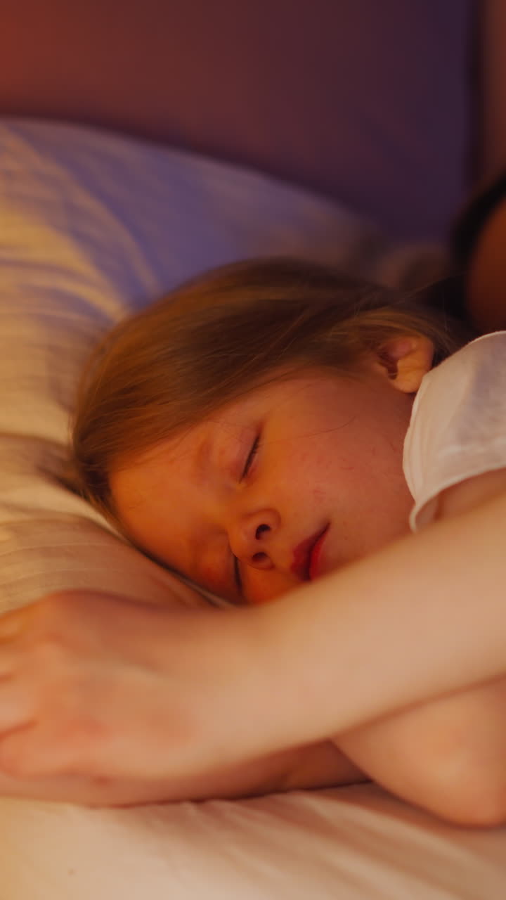 Lady hugs and strokes arm of adorable blonde daughter sleeping together in comfortable bed at orange gentle light of night lamp in bedroom closeup slow motion