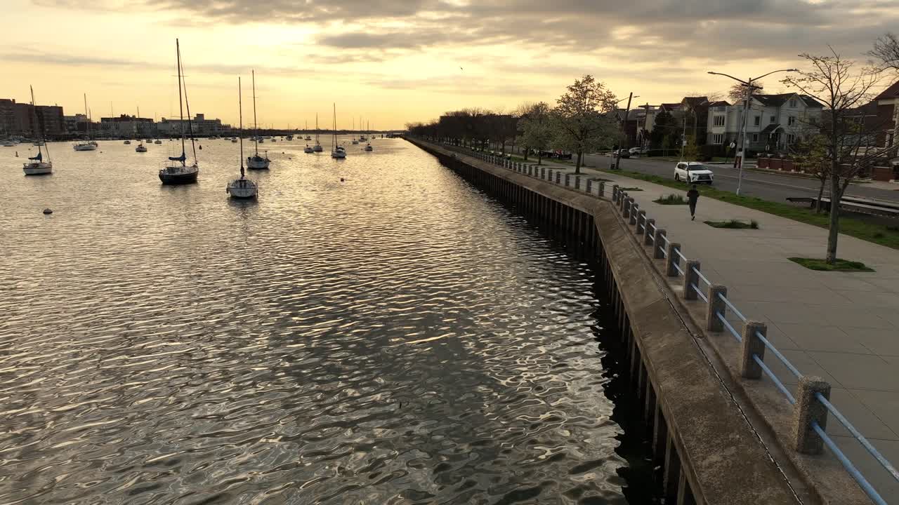 un tiro de baja altitud sobre la bahía de sheepshead durante un amanecer dorado con botes anclados y un corredor corriendo en la pasarela pavimentada a lo largo del agua
