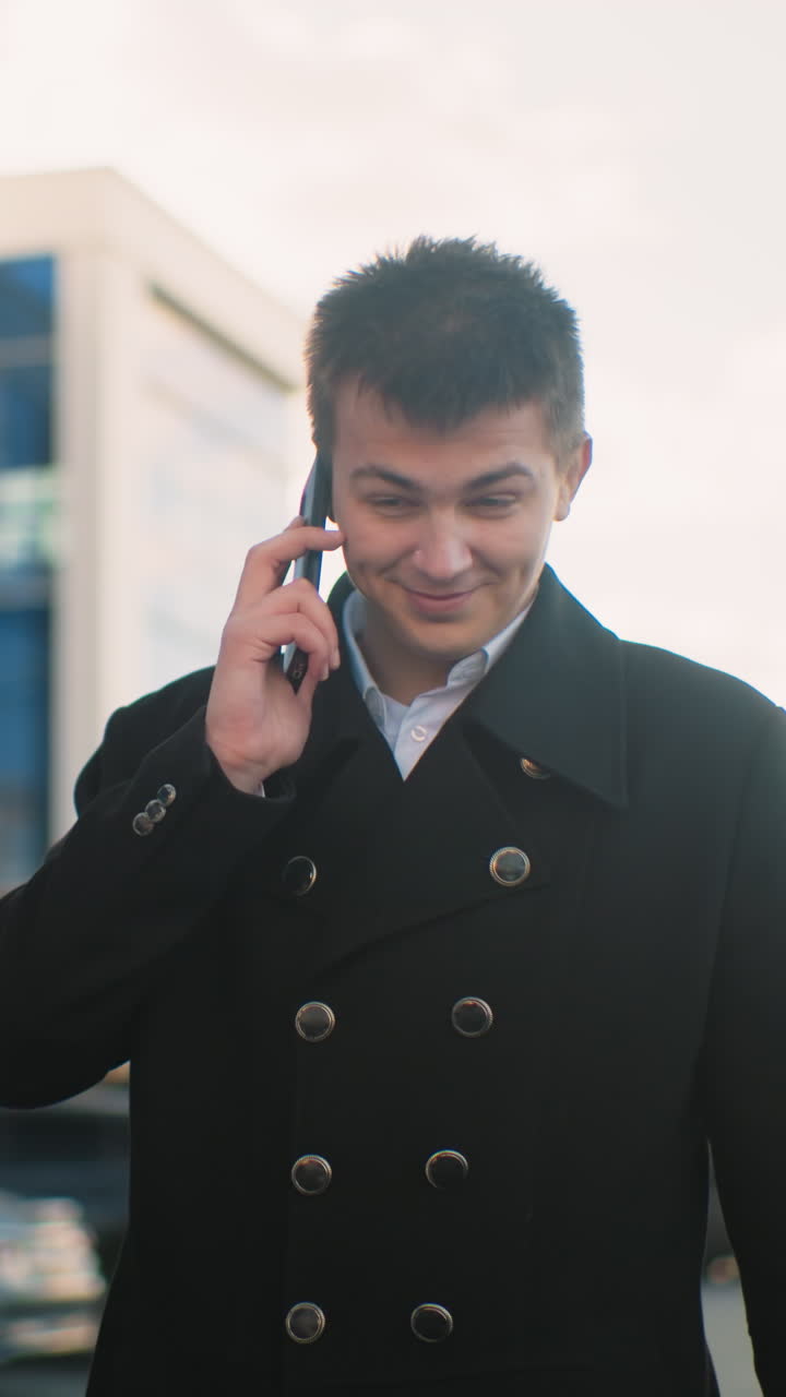 검은 코트를 입은 투자자가 흥분하며 전화로 이야기하고 현대적인 유리 건물, 부드러운 빛, 주차된 자동차, 그리고 활기찬 야외 환경을 가진 도시 지역을 가리키고 있습니다.