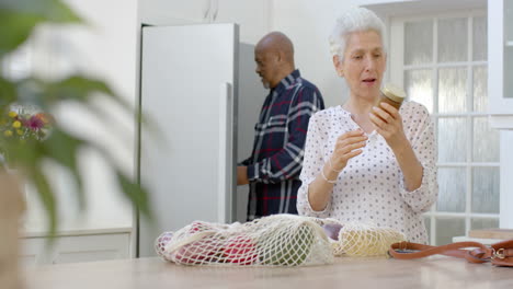 Happy-senior-biracial-couple-unpacking-shopping-bags-in-kitchen,-slow-motion