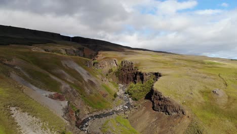 the hengifoss waterfall in iceland. the drone takes a tilt shot with the waterfall in the background and the following stream cutting through rocks. shadows of the overhead clouds can also be seen.