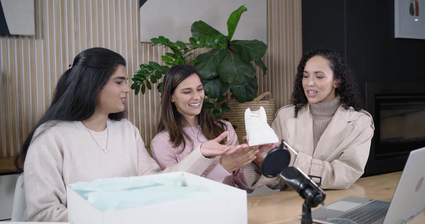 Diverse presentatrici in studio, la presentatrice di destra prende una scarpa bianca dalla scatola delle scarpe, filmando le altre che controllano il microfono
