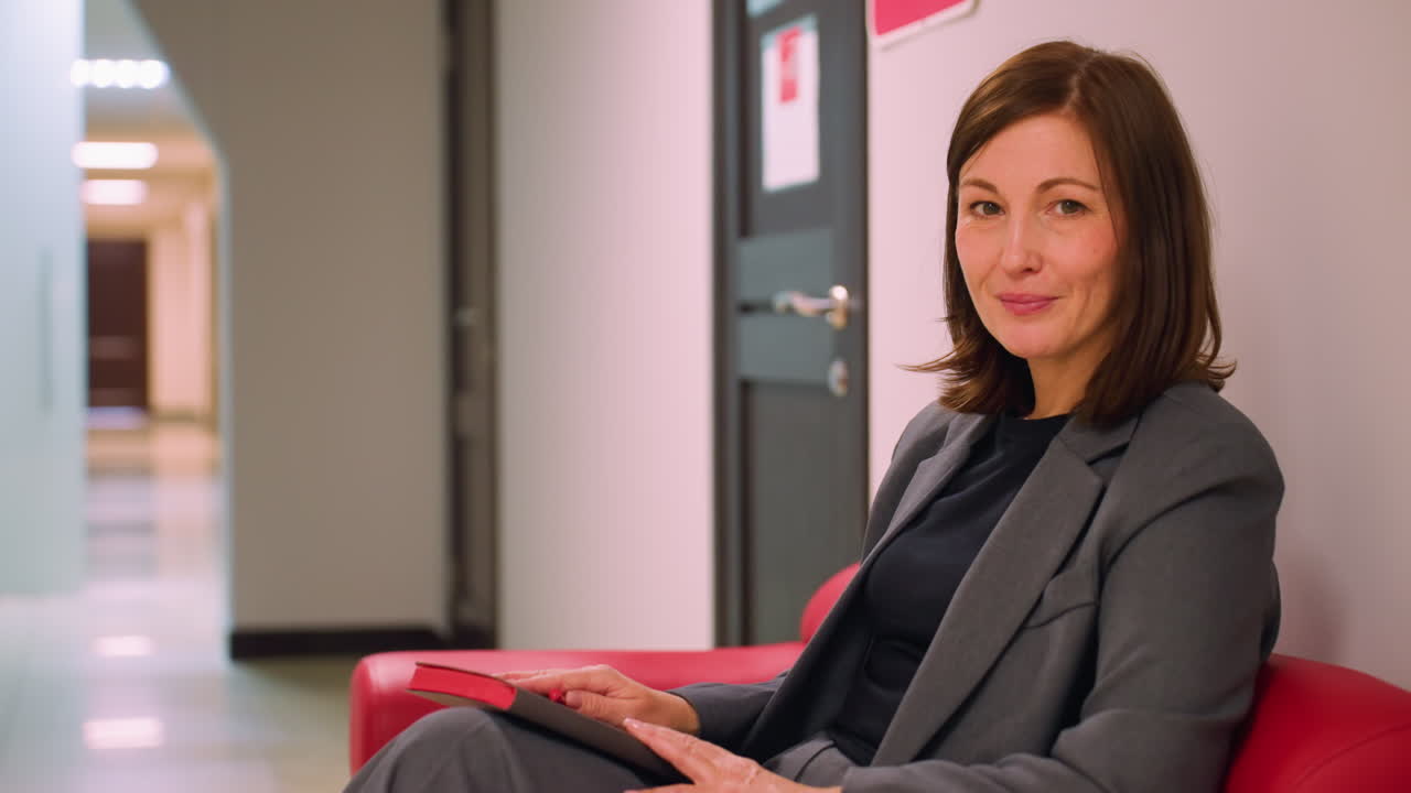 Businesswoman sitting on red couch in modern office corridor, holding tablet and looking at camera. Relaxed professional demeanor with a confident smile. Focus on modern business environment