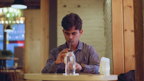 Student in formal shirt sits alone inside cozy cafe, raising coffee cup near softly lit window, with jacket on chair, water bottle on table, backpacks on bench, and warm ambient light illuminating brick walls