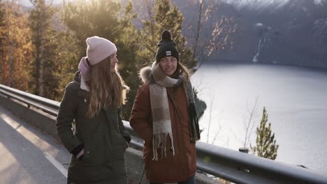 Two-female-tourists-in-winter-coats-walking-in-slow-motion-on-a-long-road-through-the-countryside-towards-the-snowy-mountain-peaks,-golden-trees-and-lake-on-the-background.-Positive-caucasian-girls-exploring-Norway-nature