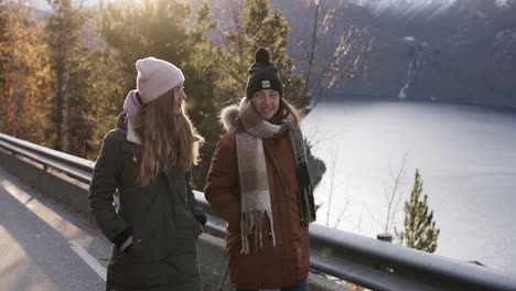 two female tourists in winter coats walking in slow motion on a long road through the countryside towards the snowy mountain peaks, golden trees and lake on the background. positive caucasian girls exploring norway nature