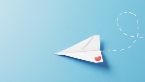 Video-of-paper-plane-with-red-heart-on-wing,-flying-on-blue-background-with-copy-space