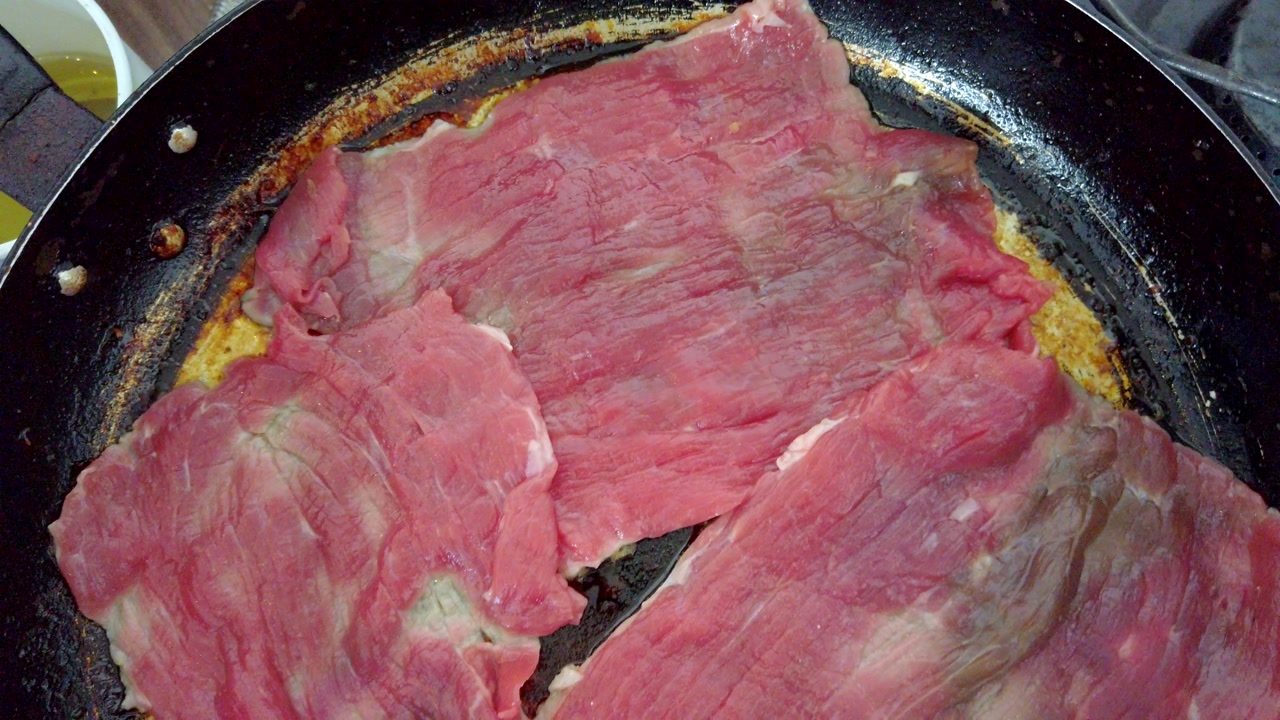 Top view of raw beef slices cooking in a frying pan on a gas stove, showing oil and kitchen tools around.
