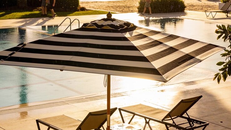 Inquadratura di un ombrellone a strisce che ombreggia due sedie a bordo piscina, due adulti che entrano da sinistra camminando per una passeggiata.