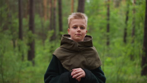 blond boy in vintage costume leans on sword in spring forest