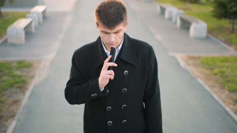 man in black coat holds phone under jaw, tapping it three times during call while appearing displeased, background includes modern glass buildings, soft sunlight, paved walkway, and urban benches