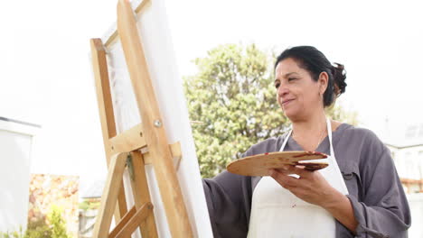 Feliz-Mujer-Mayor-Birracial-Pintando-Cuadros-Y-Sonriendo-En-Un-Jardín-Soleado,-Cámara-Lenta,-Espacio-De-Copia