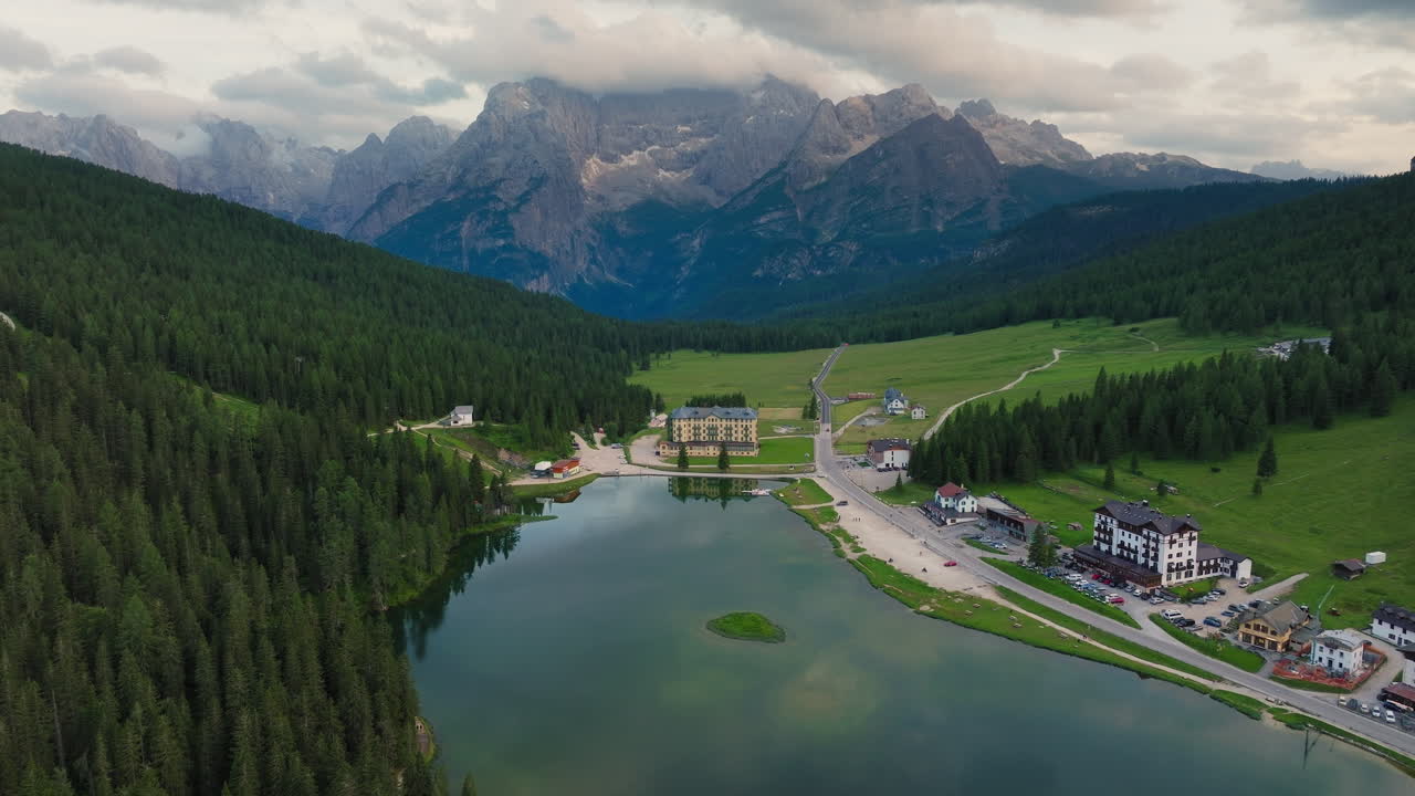 Winding road beside Lake Misurina in Dolomites with lush forest and reflective water, panoramic aerial descend