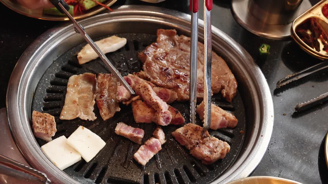 Hands use tongs and scissors to cut pork belly on Korean barbecue grill, overhead view