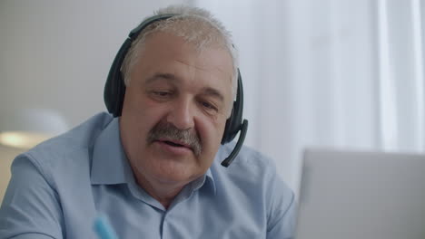 middle-aged chubby man with moustache is using headphones with microphone for communicating with colleagues at online chat