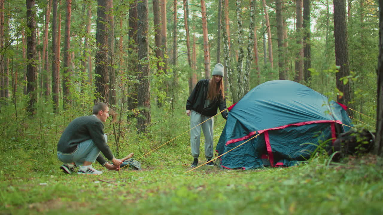 Young friends setting up camping tent in forest as woman holds yellow rope steady while man crouches to secure peg into ground surrounded by tall trees and lush greenery in calm outdoor setting
