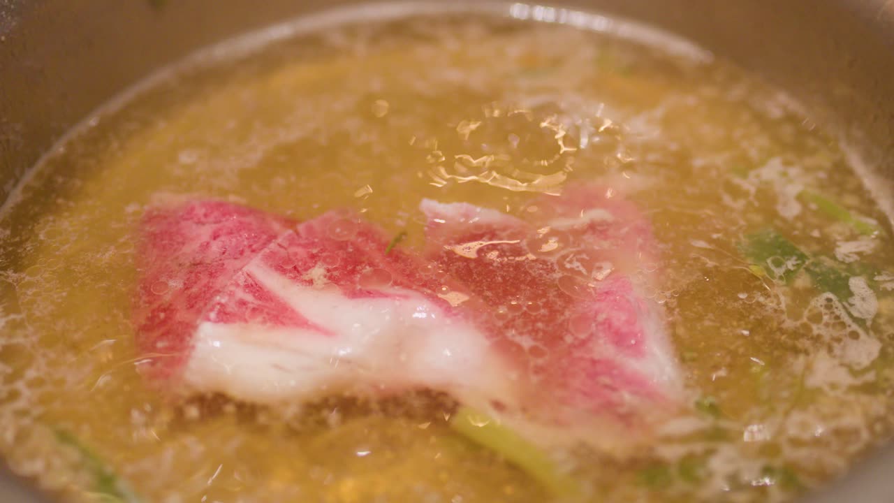 Marbled beef slices are cooked in simmering hotpot broth using chopsticks, close-up overhead view