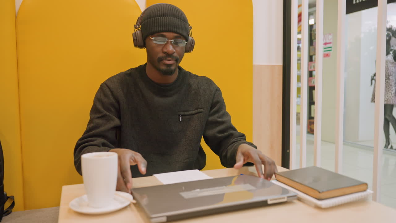 Mann lukker bærbar PC ved kafébord, gul bås som bakgrunn, hodetelefoner og kaffekopp, notatbok og penn ved siden av datamaskinen, fokusert frilanser pakker bærbar PC i ryggsekk, mykt naturlig lys og ryddig.