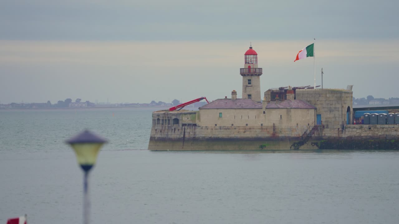 Very wide view of a 60 fps 4 k footage of a lighthouse in a Dublin harbor with the Irish flag blowing in the wind on an overcast rainy day