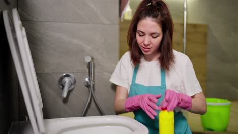 a confident brunette cleaning lady in a white t-shirt and blue apron sprays detergent from a yellow bottle on the walls of the toilet during cleaning in the bathroom in an apartment on call