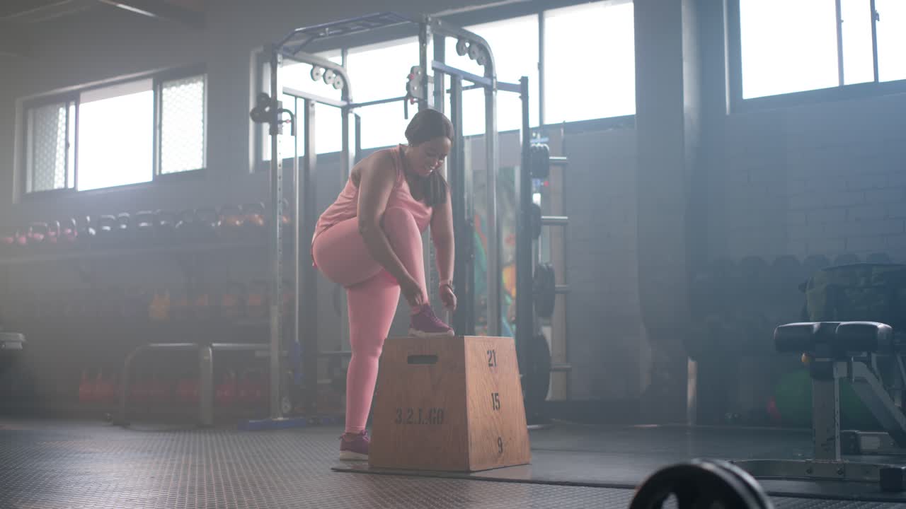 African American woman in pink approaching plyo box at gym, placing foot tying laces for step-up