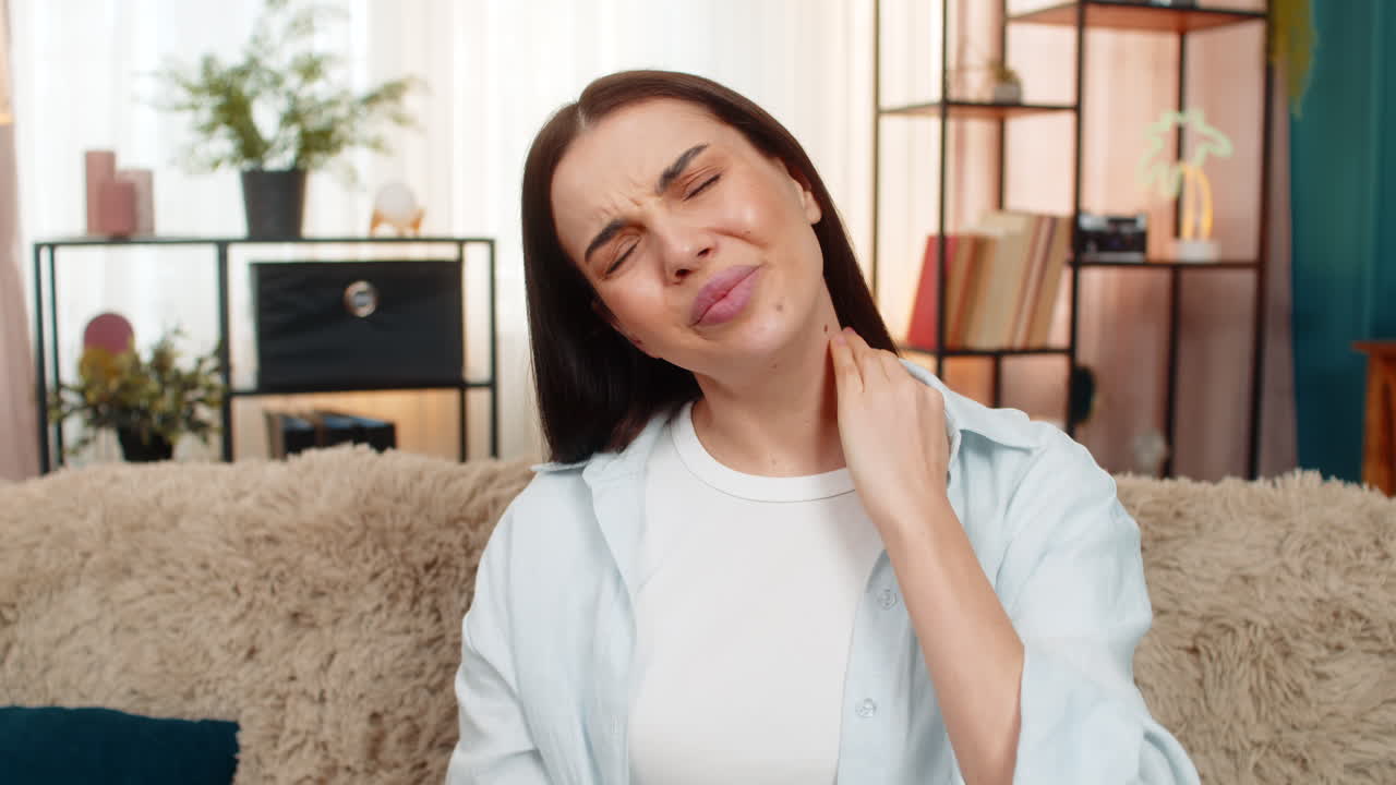 Young woman sitting on sofa holding neck feeling severe pain injury discomfort after physical trauma