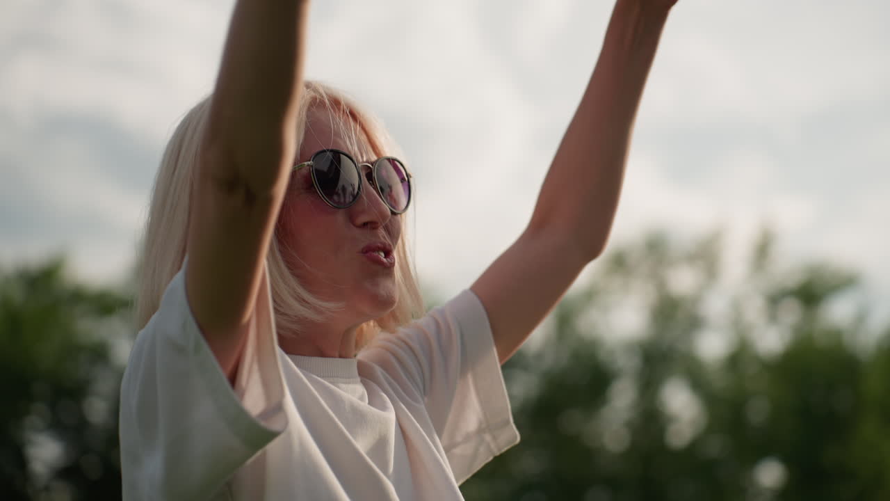 Cheerful woman wearing dark shades swaying picnic blanket overhead while smiling, sunlight filtering through trees, casual shirt, green park setting, joyful summer family preparation moment