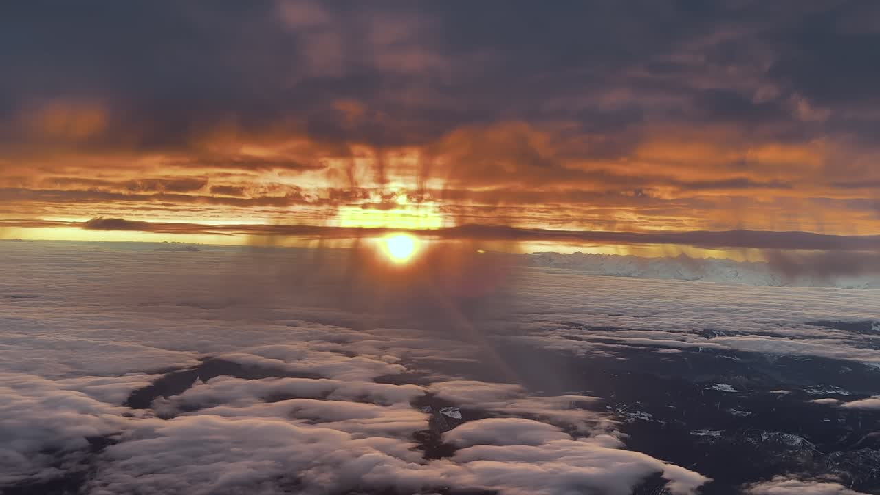 Eine Luftaufnahme eines brennenden Sonnenaufgangs über der Alpenkette, aufgenommen aus einem Jet-Cockpit, das auf Reiseflughöhe zwischen Wolkenschichten fliegt, mit einer aufgehenden Sonne am Horizont