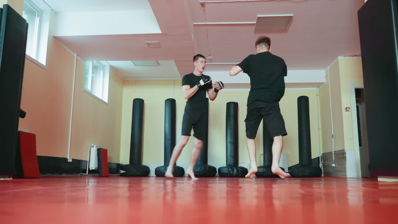 무술 체육관 안에서 태권도 수련자들이 스파링 세션을 하고 있어요. 한 명이 빠른 공격을 하는 동안 다른 한 명은 빨간색 매트 위에서 방어 자세로 막고 있고, 주변에는 펀칭백이 있으며 자연광이 창문을 통해 들어오고 있어요.