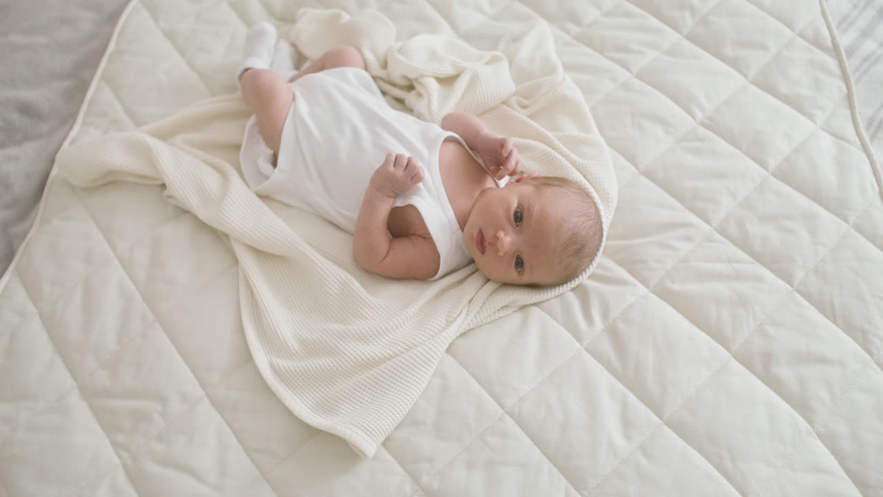 Top View Of A Baby In White Bodysuit Lying On Bed Moving His Legs And Arms