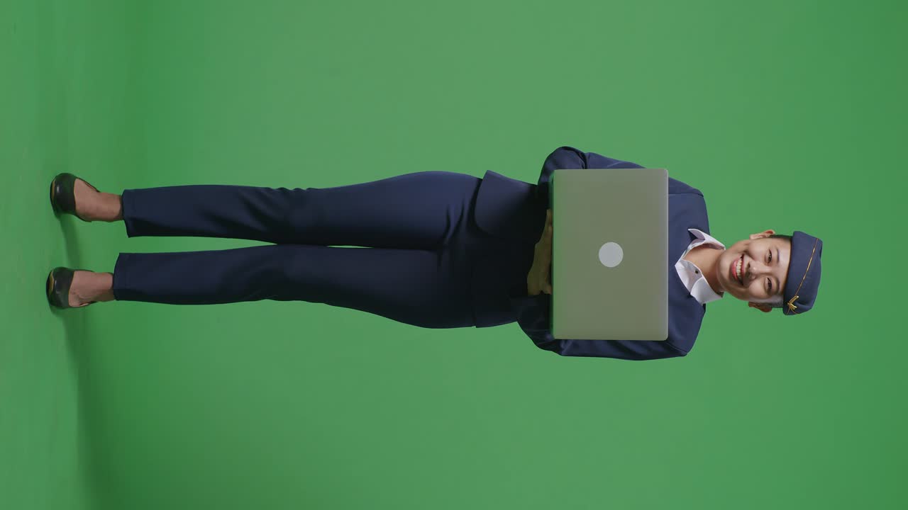 cuerpo lleno de azafata asiática usando una computadora portátil y sonriendo a la cámara mientras está de pie en el estudio de fondo de la pantalla verde