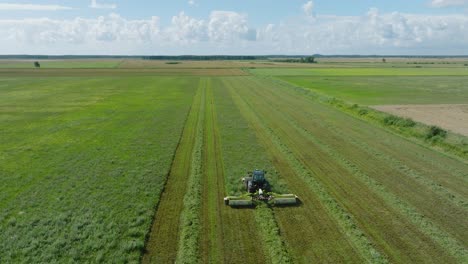 aerial establishing view of a tractor mowing a fresh green grass field, a farmer in a modern tractor preparing food for farm animals, sunny summer day, wide drone shot moving forward low