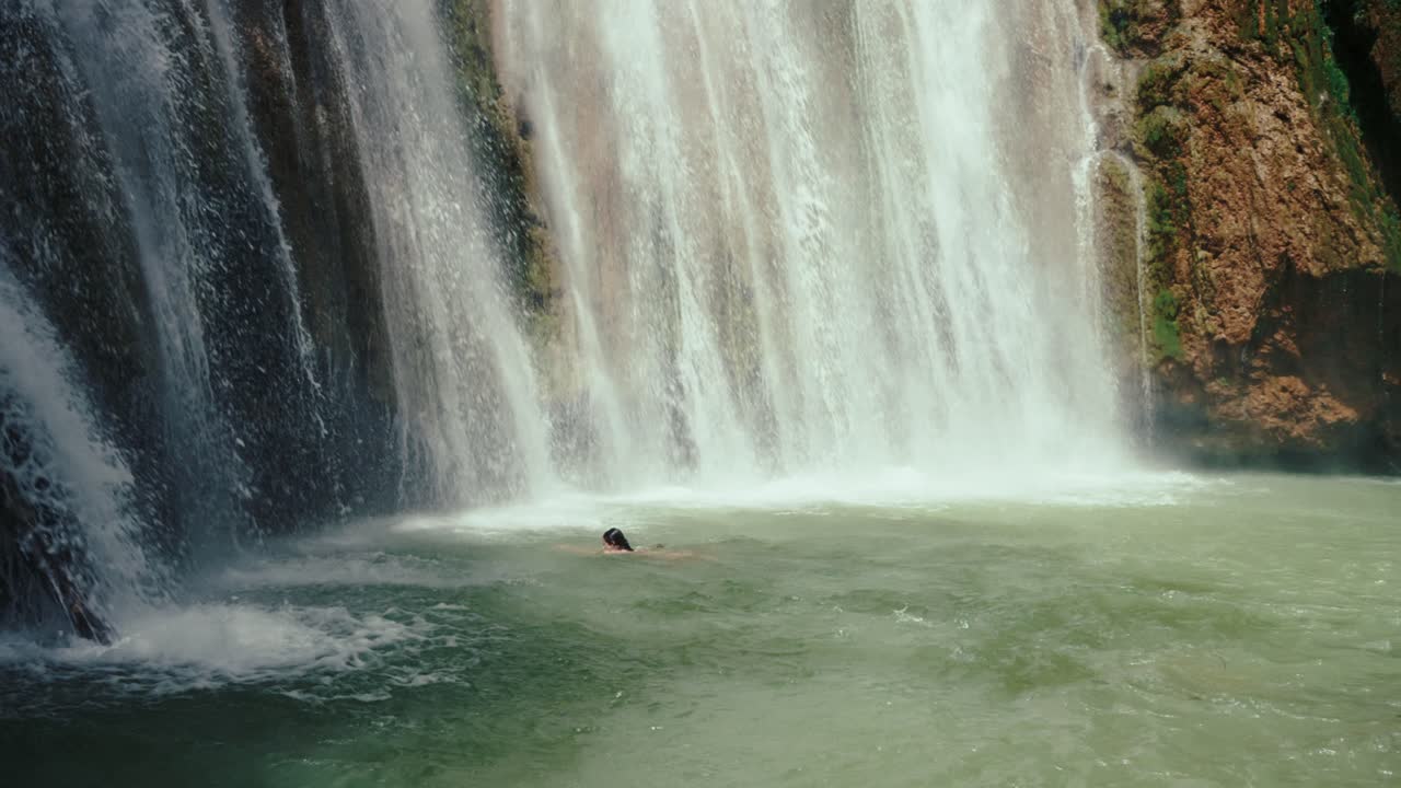 Wide composition showing a lone swimmer floating calmly beneath a roaring tropical waterfall.