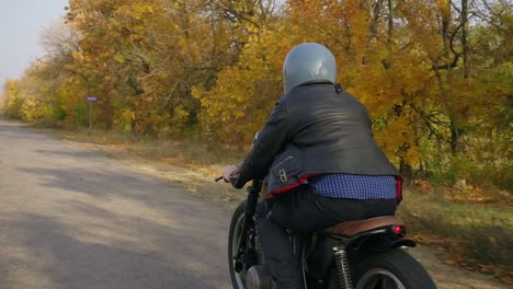 Rückansicht-Eines-Mannes-Mit-Grauem-Helm,-Lederjacke-Und-Kariertem-Hemd,-Der-An-Einem-Sonnigen-Herbsttag-Auf-Einer-Asphaltstraße-Motorrad-Fährt