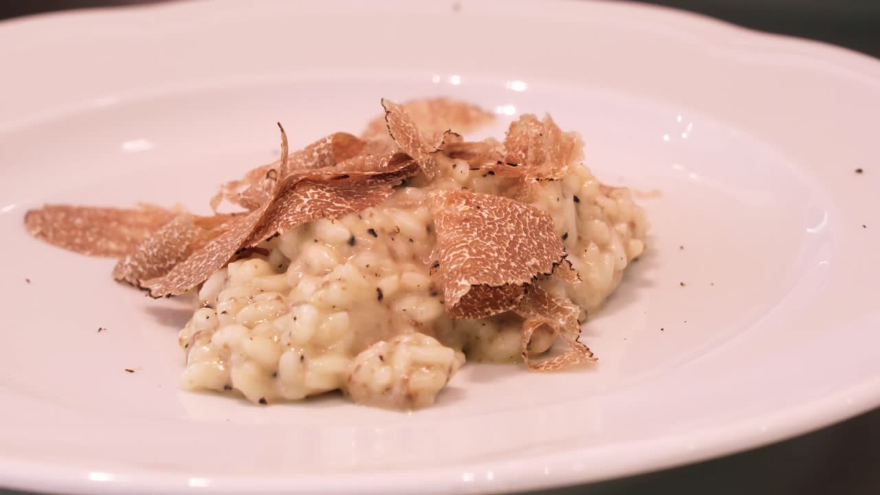 An extreme close-up shot highlights creamy risotto on a white plate, topped with thin truffle shavings, emphasizing the dish's texture and gourmet presentation.