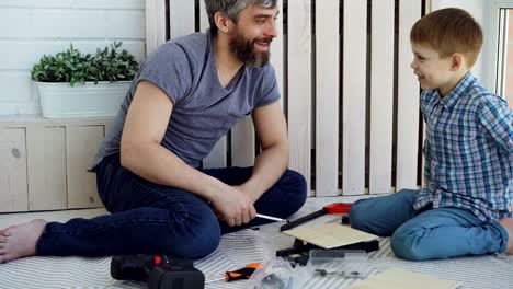 padre amistoso y hijo pequeño están hablando y riendo mientras hacen trabajo de construcción sentados en el suelo en casa. conversación, instrumentos y concepto de paternidad.