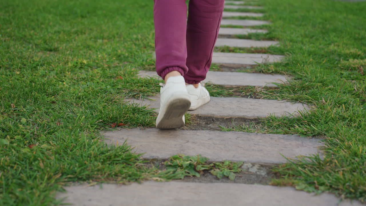Motivated woman in hoodie carrying sports bag over shoulder with hands in pocket strides along grass stepping stones toward exercise bars under blue sky in urban park setting in soft morning light