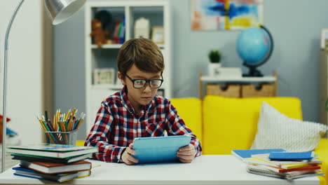 La-Cámara-Se-Acerca-Al-Pequeño-Adolescente-Con-Gafas-Y-Camisa-A-Cuadros-Mirando-Algo-En-El-Dispositivo-De-La-Tableta-En-El-Escritorio-En-La-Acogedora-Habitación-De-Los-Niños