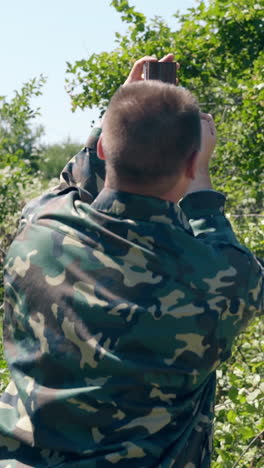 middle-aged man makes marks on metal pole top with pencil in weeds against old country house on day