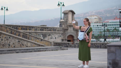 with her baby in a kangaroo backpack, a young woman captures travel memories on a mobile phone. while walking, she occasionally looks at the phone screen