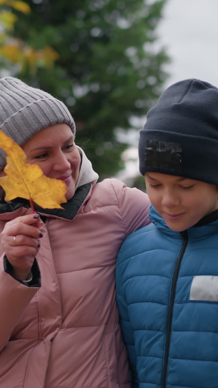 Mother in pink sweater holds son's by the shoulder and affectionately uses a leaf to touch his face, both smiling during an autumn walk